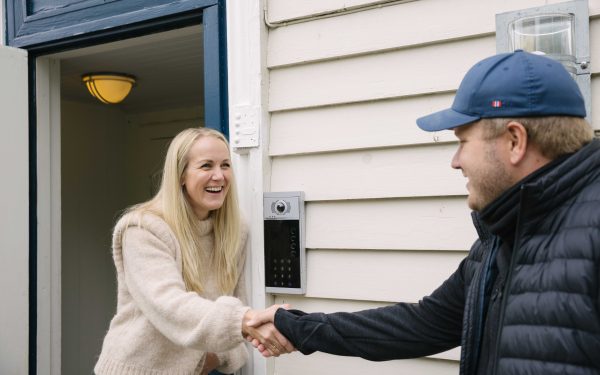 dame med blondt hår og beige ullgenser som står i døråpning og håndhilser og smiler med en elektriker i sort boblevest og blå caps som smiler, og med en porttelefon på ytterveggen av boligen